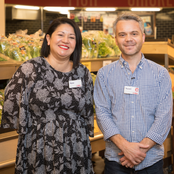 A woman in a floral dress and a man in a checked shirt smile side by side, standing in a supermarket produce aisle with name badges.